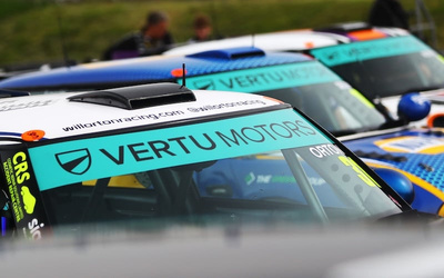 Close-up of three racing cars lined up, each featuring turquoise windscreen banners with the text 'VERTU MOTORS' and web address 'willortracing.com'. The cars have race numbers and sponsor logos visible, and the setting appears to be a racetrack with blurred figures in the background. The image highlights high-performance vehicles associated with Vertu Motors, a dealership known for new cars and used cars.
