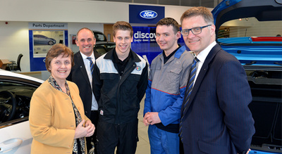 Five people standing inside a Ford car dealership, including two men in suits and three younger men wearing work overalls, with Ford branding and signage in the background near the Parts Department, showcasing a professional environment for used cars and new cars sales and servicing.