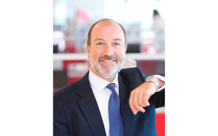Portrait of a smiling middle-aged businessman with a beard wearing a dark suit, white shirt, and blue patterned tie, seated in a bright office environment. No visible text related to used cars or new cars.