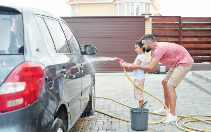 Man and young boy washing a dark blue used car in a driveway with a hose and bucket, surrounded by a residential setting. The image highlights care and maintenance of used cars, with interest in keeping both used cars and new cars clean and well-maintained.