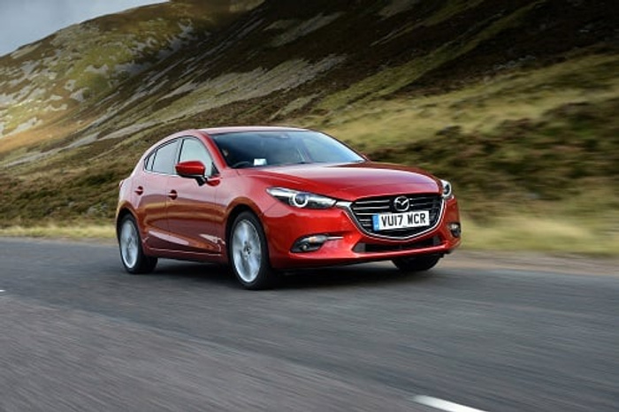 Red Mazda hatchback car driving on a winding road through a hilly landscape with grassy mountains in the background, registration plate VU17 MCR, showcasing a sleek design suitable for used cars and new cars buyers.