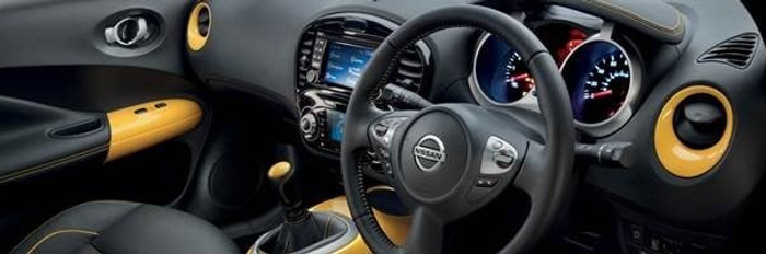 Interior view of a Nissan car showing the steering wheel with Nissan logo, dashboard with illuminated speedometer and tachometer, central touchscreen display, gear shift, and door handle. The interior features black and yellow accents. This image is relevant for used cars and new cars.
