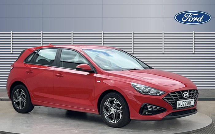 Red Hyundai hatchback car with registration plate NU72 KHT parked on a circular platform in front of a building with horizontal grey paneling and a Ford logo on the wall, representing used cars and new cars dealership.
