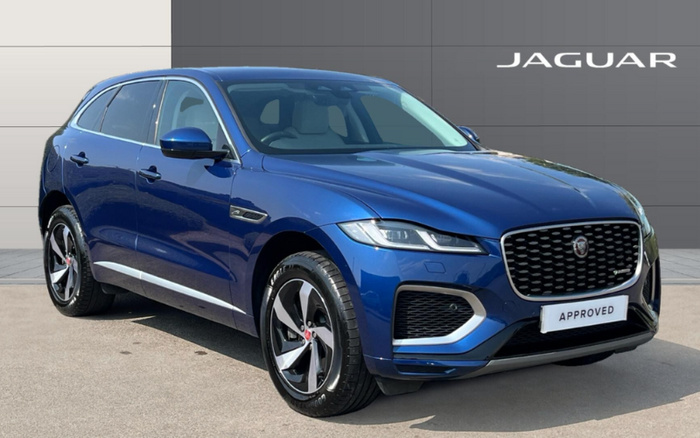 Blue Jaguar SUV parked outside a Jaguar dealership with a grey wall background displaying the Jaguar logo. The car has a front licence plate reading 'APPROVED'. This image represents both used cars and new cars available from Jaguar.
