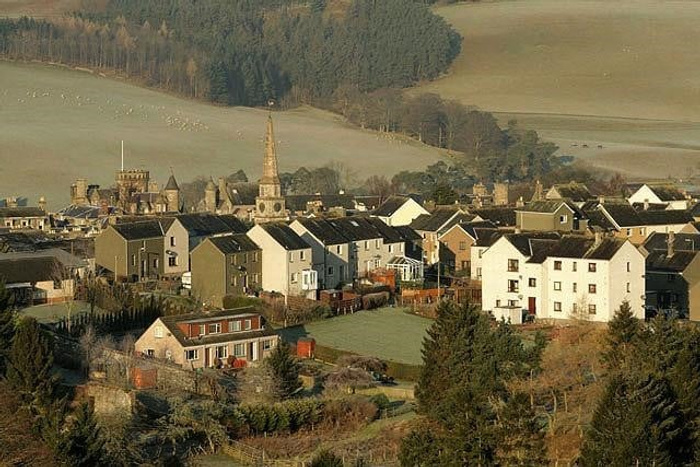 A panoramic view of a small village with densely packed houses and a prominent church with a tall spire at the centre. Surrounding the village are trees and rolling hills, with fields in the background. There are no visible cars, used cars or new cars in the image.