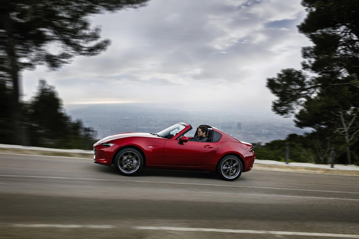 A man driving a sleek red convertible sports car on a winding road with a blurred cityscape and trees in the background, illustrating the appeal of new cars and used cars for stylish and dynamic driving experiences.