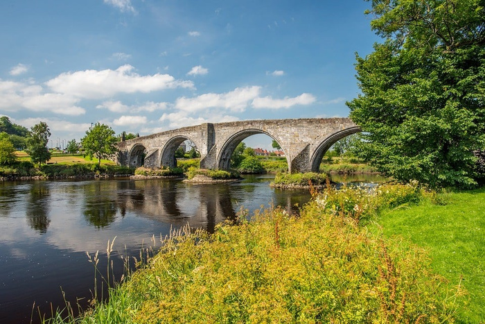 Stone bridge with multiple arches over a river surrounded by green grass and trees on a sunny day with blue sky and scattered clouds, no text visible, scene unrelated to used cars or new cars