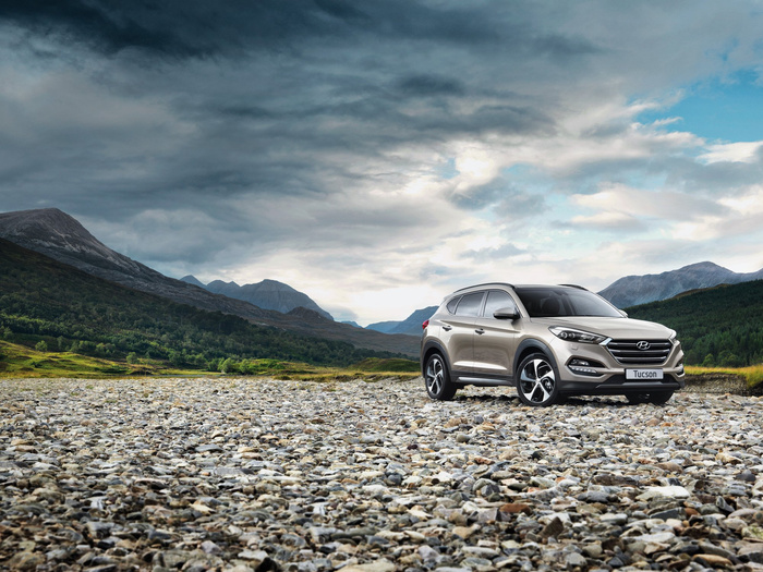 Silver Hyundai Tucson SUV parked on a rocky riverbed with mountains and dense green forest in the background under a dramatic cloudy sky with patches of blue.