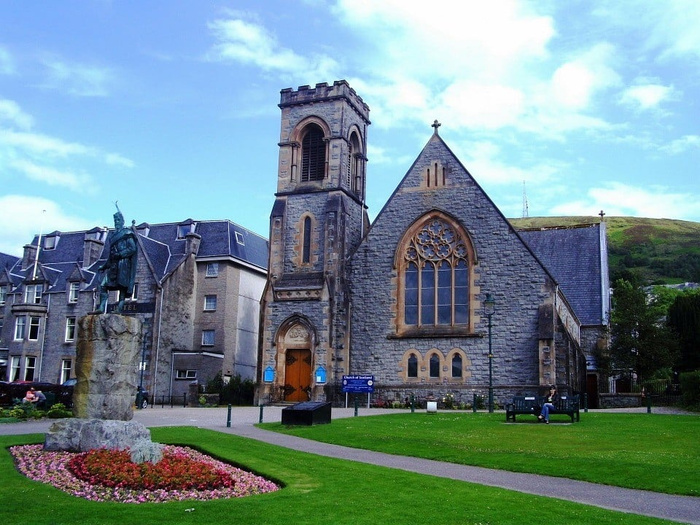 Stone church building with a tall square tower and large arched window, set in a grassy area with a paved path, a statue on a stone pedestal surrounded by flowers, and a bench where a person is sitting. No cars or text related to used cars or new cars are visible in the image.