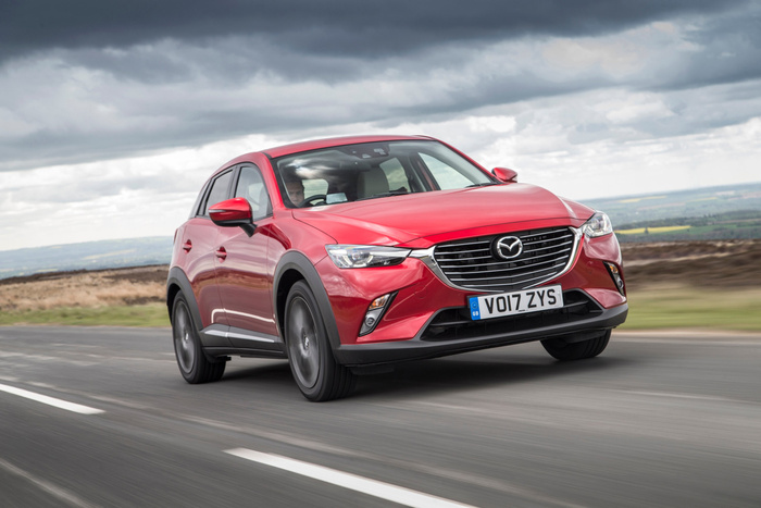 Red Mazda CX-3 SUV driving on a rural road under a cloudy sky, showing front view with visible GB registration plate VO17 ZYS, showcasing a used car or new car in motion.