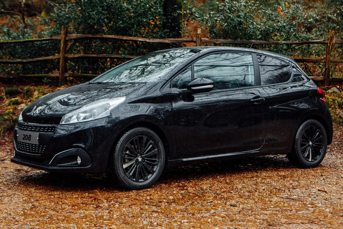Black Peugeot 208 hatchback parked on a wet gravel surface with raindrops on the car body, surrounded by autumnal foliage and a rustic wooden fence in the background, representing a quality used car or new car.