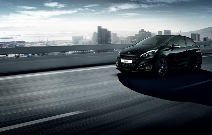 Black Peugeot 208 car driving on a modern urban highway with city buildings in the background under a partly cloudy sky, representing new cars and used cars in a dynamic setting.
