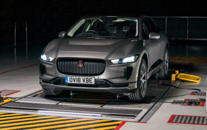 Grey Jaguar electric car with UK license plate OV18 VBE undergoing testing or inspection in a vehicle testing facility. The car is positioned on metal plates with yellow and black caution markings around it. Background is dimly lit, highlighting the vehicle's front grille and headlights. Suitable for use in promotions related to used cars and new cars.