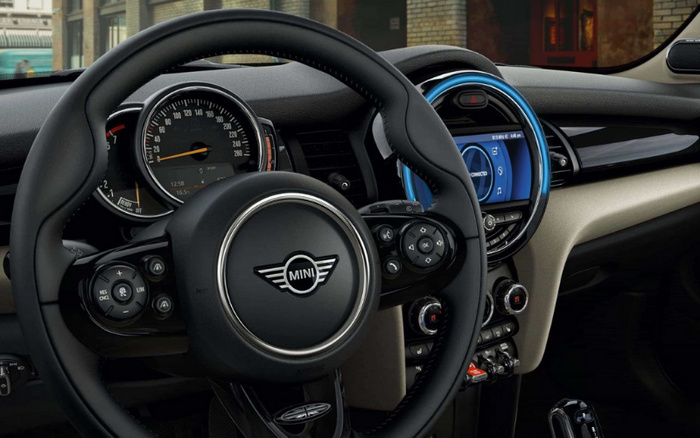 Interior view of a MINI car focusing on the steering wheel with the MINI logo, speedometer showing 0 km/h and ambient lighting around the central infotainment display. The display screen shows connectivity options and vehicle status. The dashboard features modern controls and climate settings, highlighting the stylish design of new cars, relevant for buyers interested in used cars and new cars.