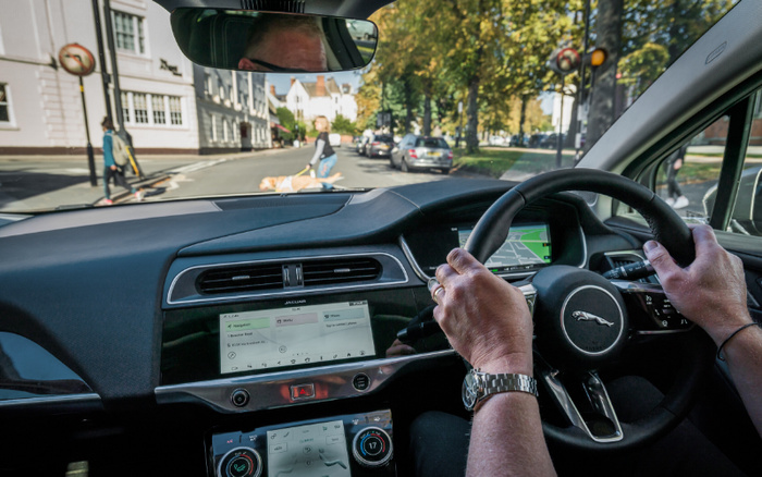 View from inside a Jaguar car showing the dashboard, steering wheel, and digital displays. The driver is holding the steering wheel with both hands and the car is navigating a street scene with pedestrians and parked cars. The dashboard screen shows radio settings with stations including Horizon, Greatest Hits Radio, and Scala Radio. This image highlights the interior of new cars, suitable for showcasing used cars and new cars for sale.