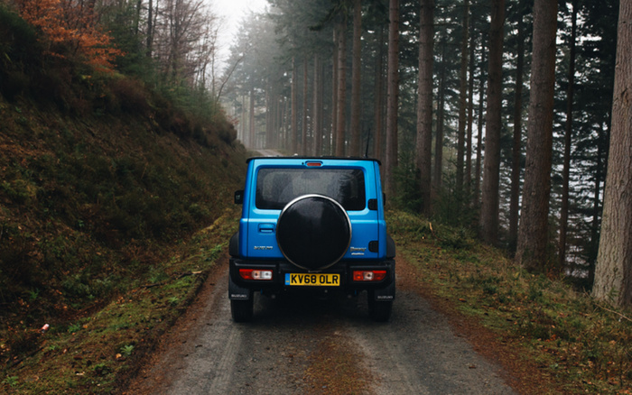 Rear view of a blue Suzuki Jimny parked on a forest gravel road surrounded by tall trees and autumn foliage. The vehicle's UK number plate reads KV68 OLR. The image evokes a sense of adventure and off-road capability, suitable for exploring new cars and used cars in rugged environments.