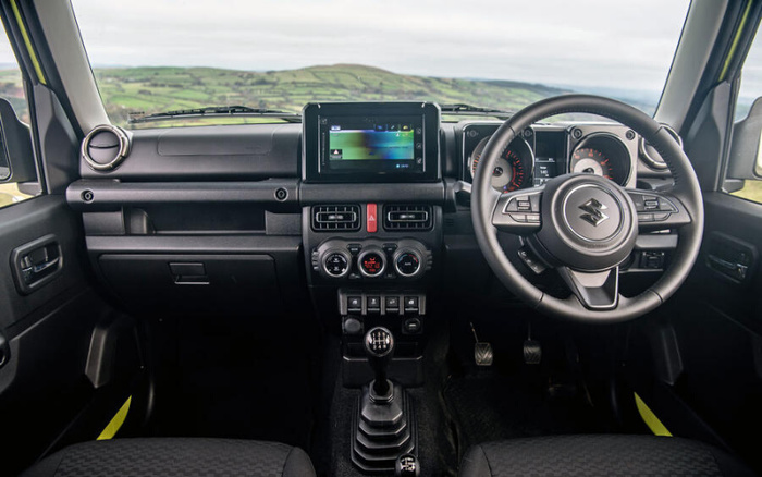 Interior view of a Suzuki car dashboard showing the steering wheel with Suzuki logo, central touchscreen display, air vents, climate control dials, and manual gear stick. The car is right-hand drive with a clear view of the countryside through the windshield. This image is relevant for used cars and new cars buying options.