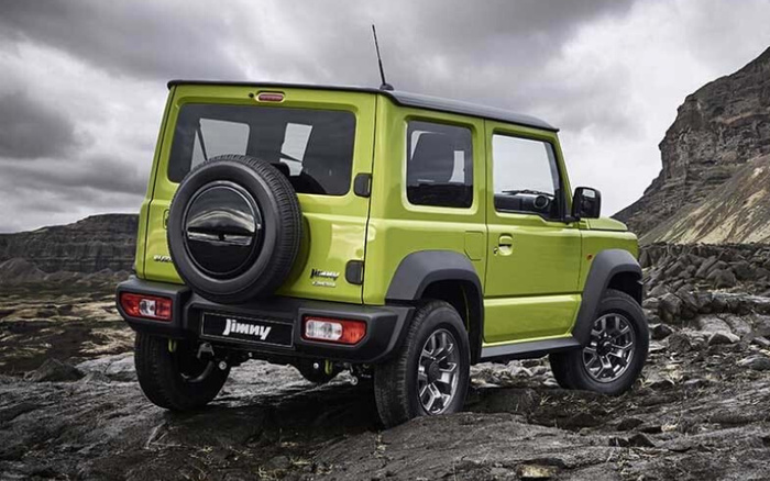 Rear and side view of a bright green Suzuki Jimny 4x4 parked on rocky terrain with dramatic cloudy sky and mountains in the background, showcasing the compact SUV's rugged design and spare wheel mounted on the rear door.