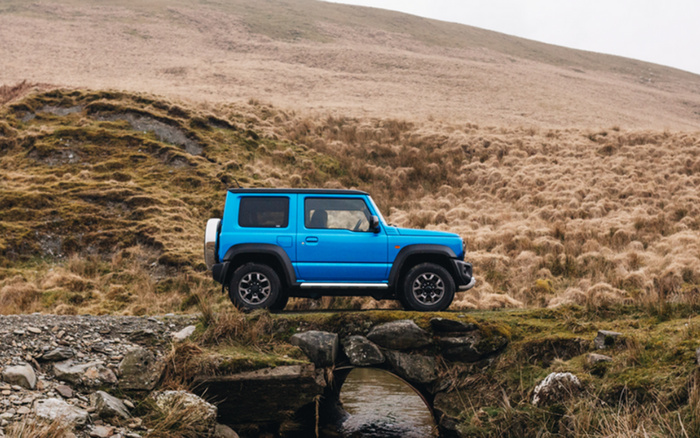 Blue off-road vehicle parked on a small stone bridge over a stream in a grassy, hilly countryside setting, representing robust used cars and new cars suitable for rugged terrain.