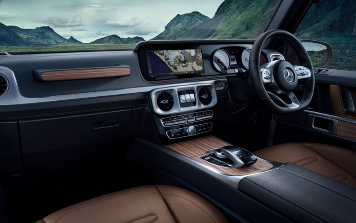 Interior view of a Mercedes-Benz car featuring a modern digital dashboard with a widescreen navigation display showing a map, circular air vents, luxurious brown leather seats, wood trim on the dashboard and centre console, and a black leather-wrapped steering wheel with Mercedes-Benz logo. The car is parked in a scenic mountainous area with green hills visible through the windows. Ideal for buyers interested in new cars and used cars.