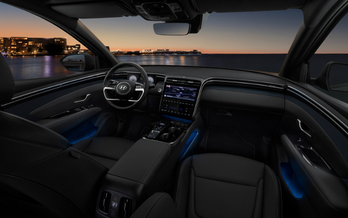 Interior view of a Hyundai car at dusk showing a sleek black dashboard, a digital instrument cluster behind the steering wheel, a large central touchscreen display with digital controls, and ambient blue lighting in the door panels. The car interior is modern and spacious with leather seats, viewed from the back seat looking towards the windshield with a waterfront cityscape in the background. The image highlights the luxury and technology features of used cars and new cars offered by Hyundai.
