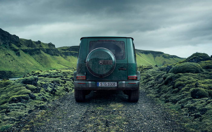 Rear view of a dark green Mercedes-Benz G-Class SUV parked on a rugged dirt path surrounded by lush green moss-covered rocks and hills under a cloudy sky, highlighting a robust vehicle ideal for adventurous drives, relevant for new cars and used cars.