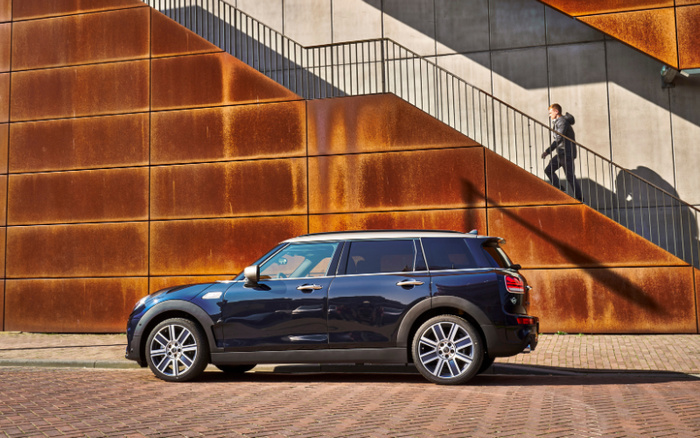 Shiny black Mini Clubman parked on a cobblestone street in front of a modern rust-coloured metal and concrete building with geometric patterns and an external staircase. A person in a black coat is walking up the stairs in the background. The image relates to used cars and new cars with a focus on the Mini brand vehicle.