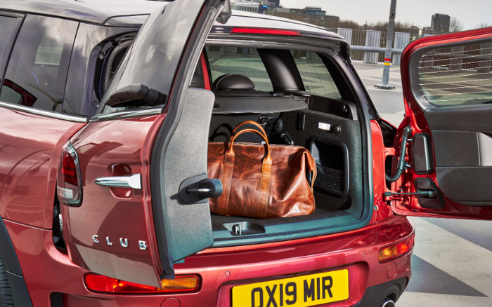 Open rear view of a red MINI Clubman showing the split rear doors with one door swung open to the side and the other open upwards, revealing a brown leather travel bag inside the spacious boot. The car is parked on a rooftop parking lot with city buildings in the background. The car's registration plate reads OX19 MIR.