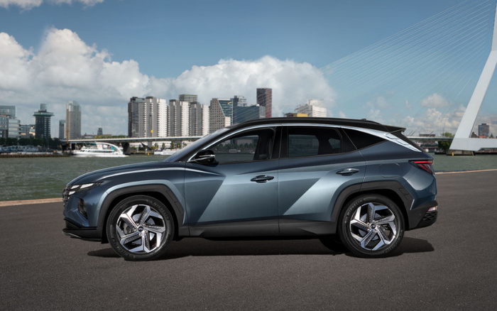 Side view of a modern blue-grey SUV parked near a riverfront with a city skyline and a large white suspension bridge in the background. The car is a new model with sleek, angular design and shiny alloy wheels. Clear blue sky with scattered clouds enhances the urban setting. Ideal image for showcasing new cars and used cars in a city environment.