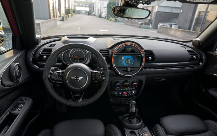 Interior view of a modern Mini Cooper car showcasing the dashboard with a circular central digital display screen, steering wheel with multiple control buttons, speedometer showing 0 mph, climate control knobs, gear shift, and black leather seats. The scene is taken from the driver's perspective looking out through the windshield onto a city street. This image is suitable for advertising used cars and new cars.