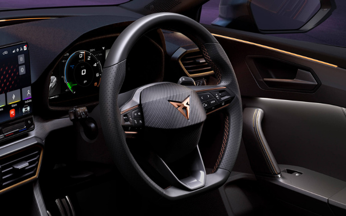 Close-up view of a modern car interior featuring a carbon fibre-textured steering wheel with a copper-coloured Cupra logo at the centre. The dashboard includes a digital speedometer display showing zero speed and gear in 'P', alongside a touchscreen control panel with various menu icons. The interior showcases premium materials with orange stitching detail on the steering wheel and door panel, highlighting the design of both used cars and new cars.