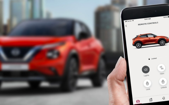 Hand holding smartphone displaying car remote controls app with an orange crossover SUV shown on screen, options for Lights, Horn, Lock, and Unlock, status showing the car as Locked. In the blurry background, a matching orange crossover SUV is parked outdoors, with urban buildings visible behind it. Keywords include used cars and new cars.