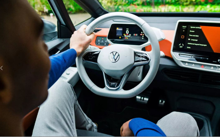 Interior view of a person sitting in the driver's seat of a Volkswagen electric car, holding the white steering wheel with the VW logo. The dashboard displays digital speed and various indicators, while the centre touchscreen shows system settings including screen, time and date, language, additional keyboard languages, and units. The modern car interior has a clean design with orange accents, highlighting features relevant for both used cars and new cars buyers.