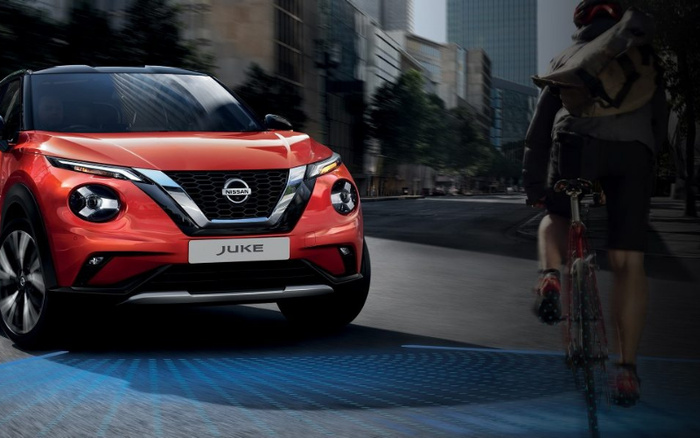 Front view of a red Nissan Juke car driving on a city street with skyscrapers in the background, next to a cyclist wearing a backpack. The Nissan badge and model name 'JUKE' are clearly visible on the front grille and licence plate. The scene highlights modern urban transport, suitable for advertising used cars and new cars.