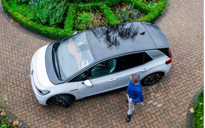Aerial view of a man in a blue sweater and grey trousers standing next to a silver and black compact hatchback car parked on a paved driveway bordered by green shrubs. The car is a modern new car with sleek design details, captured in a residential garden setting, representing options for used cars and new cars.