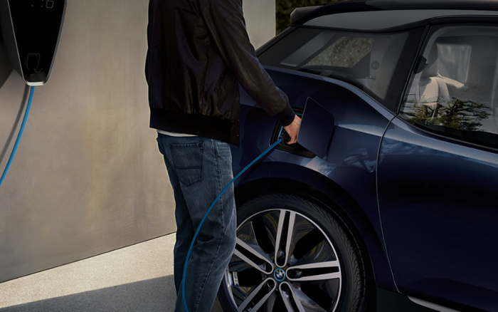 Person charging a dark blue BMW electric car using a blue charging cable connected to a wall-mounted charger. The image highlights the rear side of the car and emphasises eco-friendly new cars, with relevance to used cars and new cars market options.