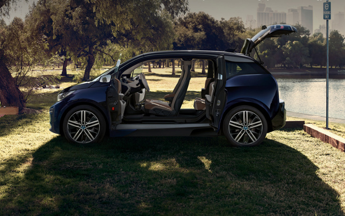 Side view of a modern black compact electric car with all doors and rear hatch open, parked on grass near a lake with trees and city skyline in the background, under natural daylight, illustrating a spacious interior and innovative door design, suitable for used cars and new cars buyers.