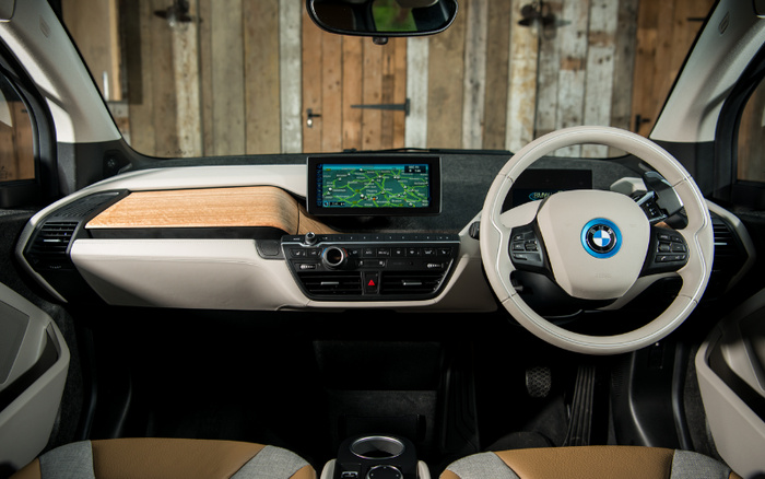 Interior view of a BMW car showing the dashboard and steering wheel. The dashboard features a central digital display showing a navigation map with locations and routes. The steering wheel has the BMW logo in the centre and multiple control buttons. The interior design includes a combination of beige and brown colours with wooden accents on the dashboard. This image is relevant for used cars and new cars, showcasing modern car technology and stylish design.