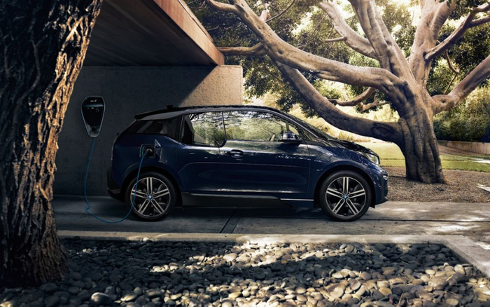 Dark blue electric car charging under a wooden overhang in a serene garden setting with large trees and stones. The vehicle is plugged into a wall-mounted charger. This scene highlights modern new cars with electric charging capabilities, appealing to buyers interested in new and used cars with eco-friendly features.