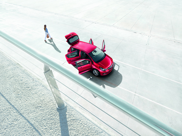 A bright red car with all doors and trunk open parked on a large, empty concrete surface, viewed from above. A person in a white dress is walking away from the car. The scene evokes a spacious, modern environment suitable for viewing used cars or new cars.