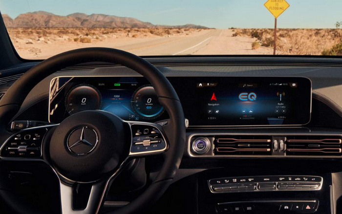 Interior view of a Mercedes-Benz car dashboard and steering wheel showing digital instrument cluster and central touchscreen display with EQ and navigation interface. The car is stationary on a desert road with a yellow road sign reading 'SUBJECT TO FLOODING' visible through the windshield. Optimised for used cars and new cars.