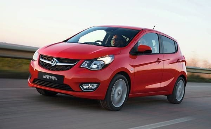 Red Vauxhall New Viva compact car driving on a road at dusk with a male driver wearing sunglasses, showcasing a new model ideal for used cars and new cars buyers seeking a small, stylish vehicle.