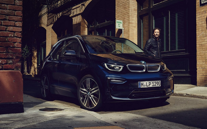Dark blue BMW i3 electric car parked on a city street with a man standing nearby wearing a black leather jacket. The car has a German license plate reading 'M LP 129G'. The background shows brick buildings with windows and shutters. A green sign on a pillar says 'PARKING ANY TIME'. The image represents used cars and new cars in an urban setting.