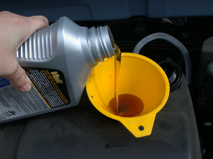 A close-up of a hand pouring engine oil from a silver bottle into a yellow funnel placed in a car engine. The bottle has a partially visible label with the word 'Visit' and safety information. This image relates to car maintenance, relevant for users interested in used cars and new cars upkeep.