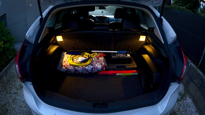 Open boot of a white hatchback car with interior lights on, showing a folded patterned blanket, yellow jumper cables, a black battery pack with white text and a red emergency warning triangle in yellow packaging labelled 'Euro Warning Pack'. The image is suitable for used cars and new cars cargo space demonstration.