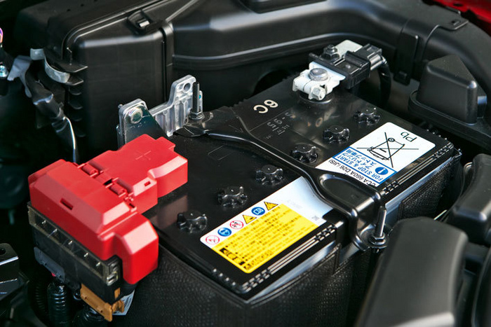Close-up of a car battery under the bonnet, showcasing the positive terminal with a red cover and safety warning labels including a yellow caution label and a white label indicating recycling instructions and battery specifications. Suitable for used cars and new cars maintenance and battery replacement.