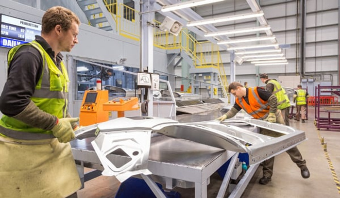 Two factory workers in high-visibility vests inspecting and working on a large metal car body panel inside a spacious automotive manufacturing plant with industrial equipment and production signage in the background, representing the production process of new cars and quality control relevant to the used cars and new cars industry.
