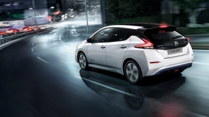 White Nissan Leaf electric car driving on a wet urban road at night, with the rear badge showing 'LEAF' and 'Zero Emission' text. The image highlights the sleek, modern design of the vehicle, suitable for advertising used cars and new cars.