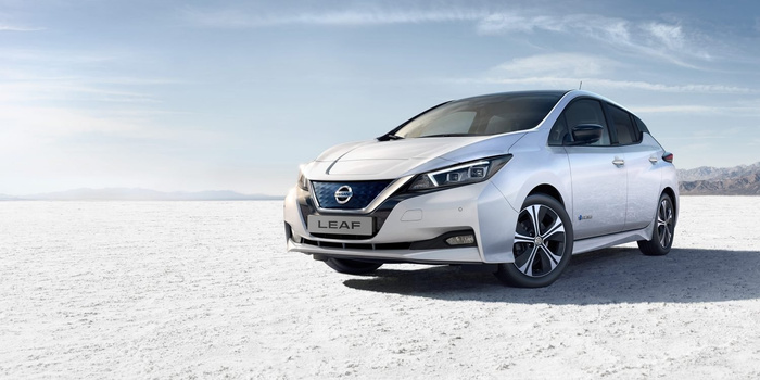 Silver Nissan LEAF electric hatchback car parked on a vast white salt flat with mountains in the background and a partly cloudy blue sky. Front license plate reads LEAF and the Nissan logo is prominently displayed on the grille. The car features sleek headlights, black and silver alloy wheels, and tinted windows. Ideal image for used cars and new cars listings or advertisements.