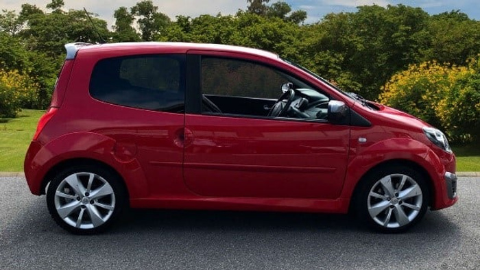 Side view of a shiny red compact car parked on a road with green trees and bushes in the background, suitable for advertising used cars or new cars.